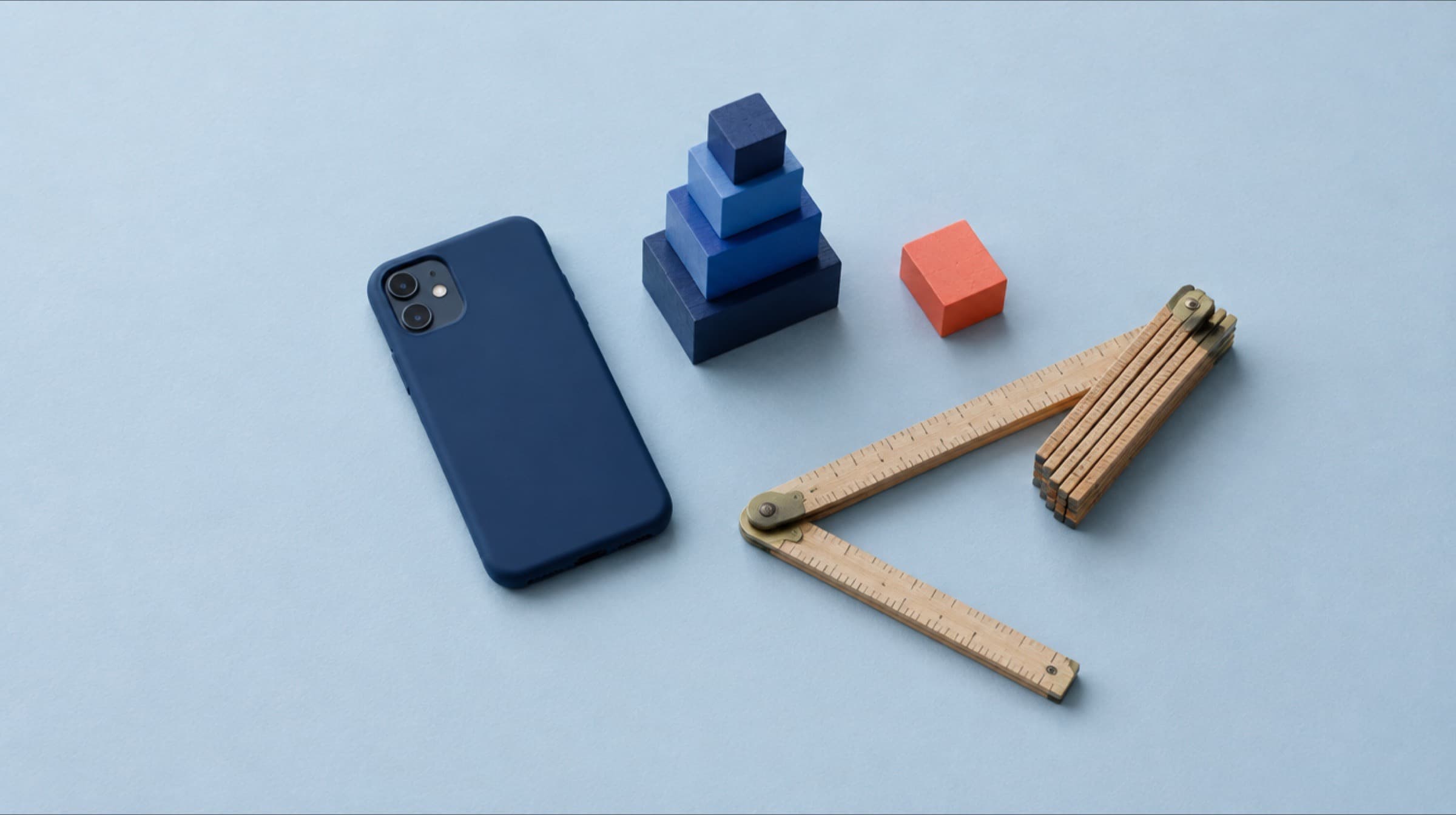 A phone in a navy case face down on pale blue paper beside a stack of navy and cobalt wooden blocks of different sizes, a folding wooden ruler and one coral block set apart