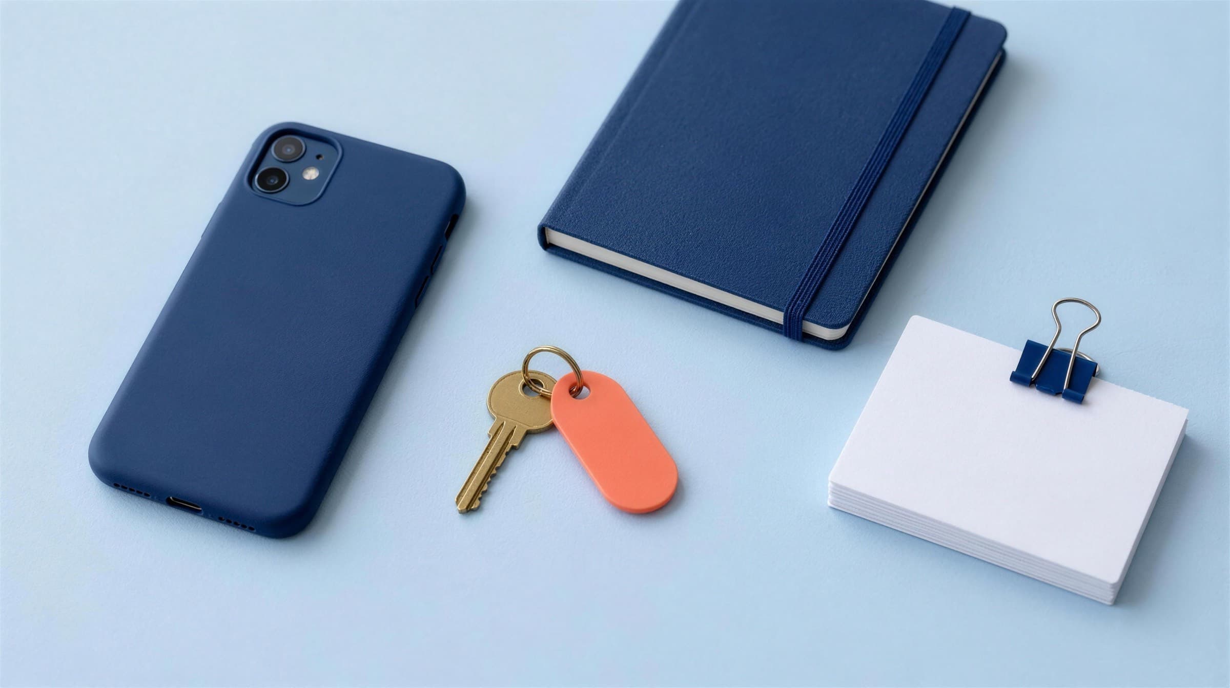 A phone in a navy case face down on pale blue paper beside a brass key on a coral tag, a navy notebook with an elastic band and a stack of blank cards in a navy binder clip
