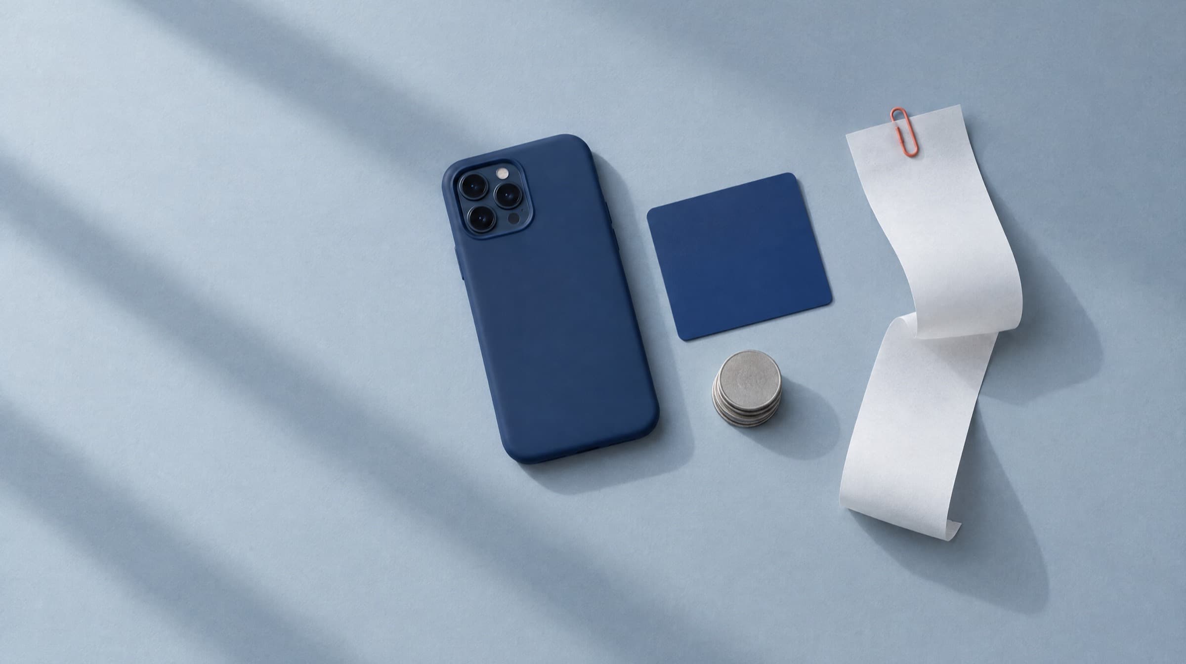 A phone face down in a navy case on pale blue paper beside a plain navy card, a short stack of coins and a curled blank receipt held by a coral paper clip